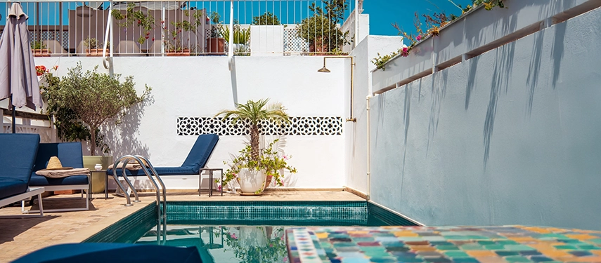 Inviting swimming pool area with sun loungers, blue cushions, and vibrant greenery against whitewashed walls.