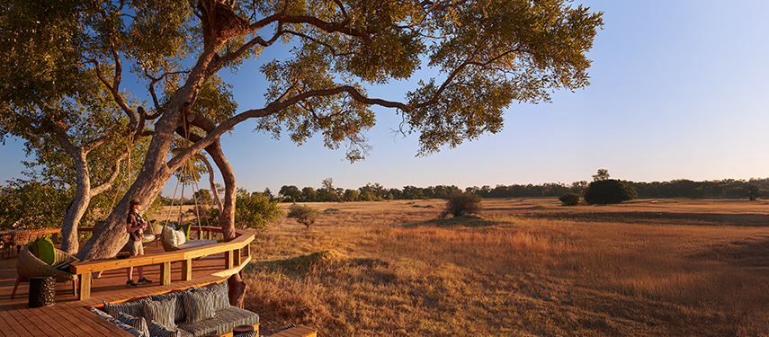 Elevated wooden deck at Tuludi Camp overlooking golden plains, with a guest relaxing under a shady tree at dusk.