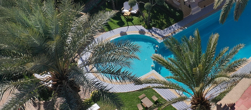 Expansive view of Ksar El Kabbaba’s kasbah architecture with tall towers overlooking the pool and gardens. Aerial view of Ksar El Kabbaba showing the kasbah’s towers, lush palm gardens, and central swimming pool courtyard.