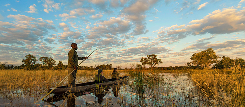 Guests enjoying a traditional mokoro canoe safari through shallow waters and golden grasses under a pastel sunrise sky.