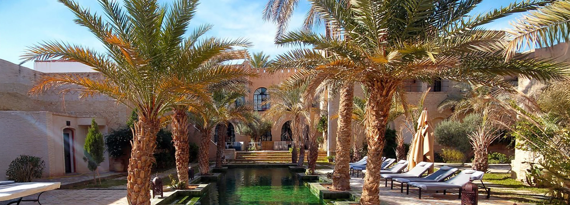 Palm-lined courtyard pool at Dar Tozeur with sun loungers, lanterns, and traditional architecture under a bright blue sky.