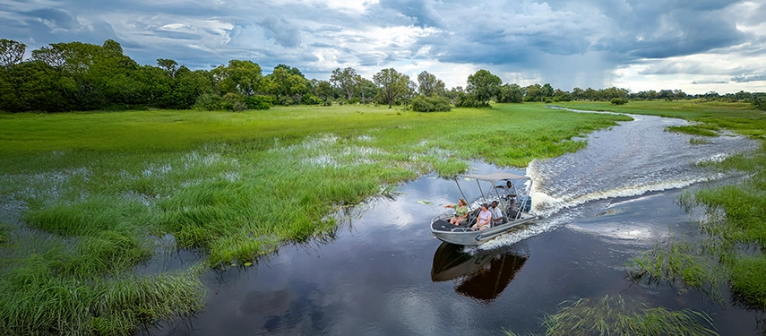 A motorboat glides along a winding Okavango channel bordered by emerald grasses under a dramatic sky.