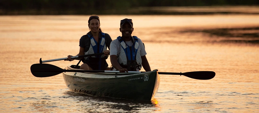 Two guests paddle a canoe across calm waters at sunset, exploring Gorongosa’s landscapes by river as golden light reflects on the surface.