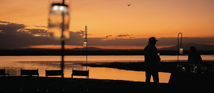 Silhouetted figures enjoy sundowners beside a tranquil lake at dusk, lanterns glowing softly against Gorongosa’s sunset horizon.