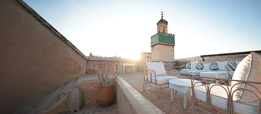Rooftop terrace at Dar Bensouda with white lounge chairs and a view of a traditional minaret at sunset.