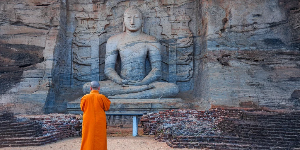 Buddhist monk in orange robes gazing up at an ancient rock-carved Buddha statue in Sri Lanka.