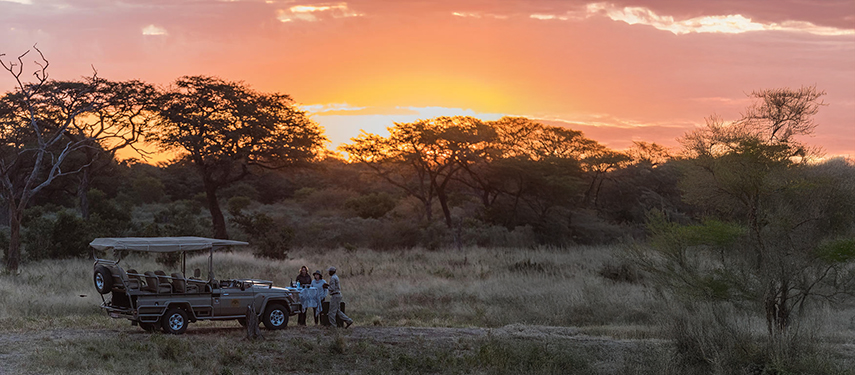 Guests stand beside a game drive vehicle during a sunset stop in a savannah landscape.