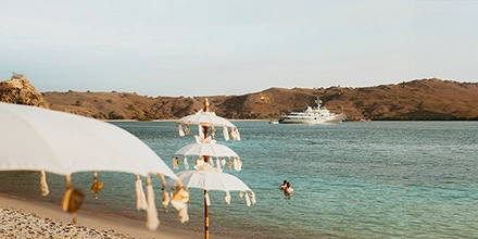 Elegant beach setup with white parasols and soft sand, Aqua Blu anchored offshore beneath warm afternoon light.