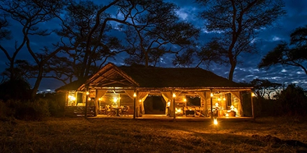 A softly lit open-air mess area glows beneath towering acacia silhouettes as dusk settles over the Tarangire wilderness.