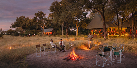 Guests relax around a firepit at Mbamba Camp as the sun sets over the Okavango Delta.