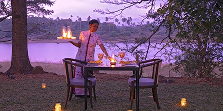 A lakeside dining table at Jetwing Kandy Gallery is set beneath soft twilight as a staff member presents flaming dishes beside the water.