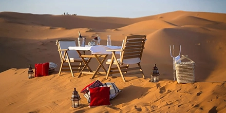 Romantic desert dining scene with table, chairs, lanterns, and red cushions on the sand dunes.
