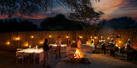 Evening boma setting with chefs preparing meals beside a glowing fire and lantern-lit tables.