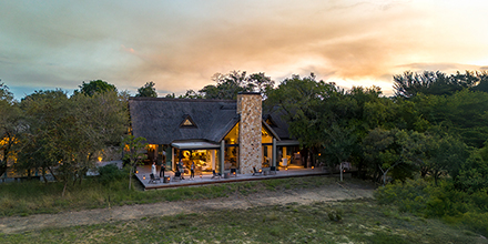A view of Monwana Lodge at sunset, with glowing interiors and a thatched roof surrounded by bushland.