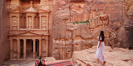 A woman in a flowing white dress stands on a rocky ledge overlooking Petra’s iconic Treasury, framed by towering rose-coloured sandstone cliffs.