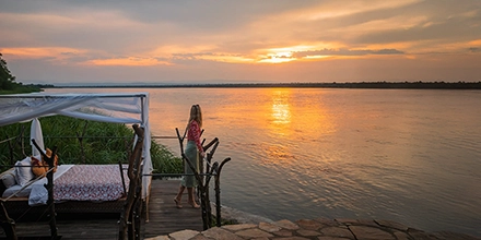 A guest stands on a wooden deck at sunset, gazing across the wide Victoria Nile as the sky glows with warm evening light.