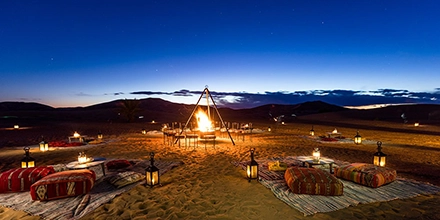 Cosy desert campfire set with lanterns and cushions glowing under the evening sky at Riad Madu Luxury Desert Camp.
