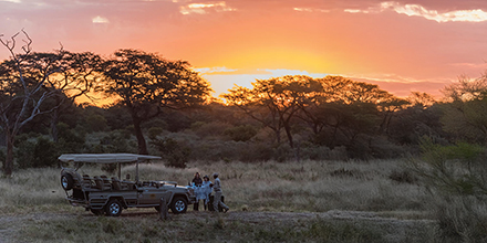 Guests enjoy a sundowner beside a safari vehicle with a vivid orange sunset over the Hwange landscape.