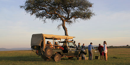 A game vehicle parked beneath a tree with guests enjoying sundowners on Kenya's Masai Mara