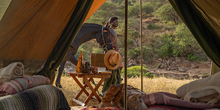 View from inside a fly tent showing a Samburu warrior with a horse, alongside rustic furnishings and layered blankets.