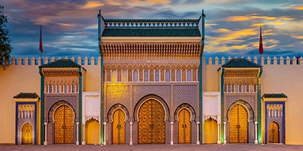 Royal Palace of Fès with golden gates, intricate mosaic arches, and green tiled roofs illuminated at dusk.