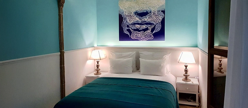 A stylish bedroom with turquoise walls, contemporary artwork, and a neatly made bed with deep blue bedding.