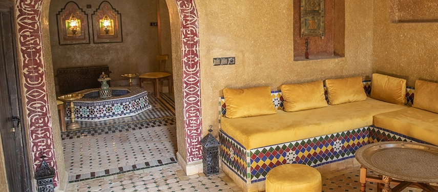 Warm Moroccan lounge with yellow cushioned seating, mosaic details, and carved archways leading to a traditional sitting area.