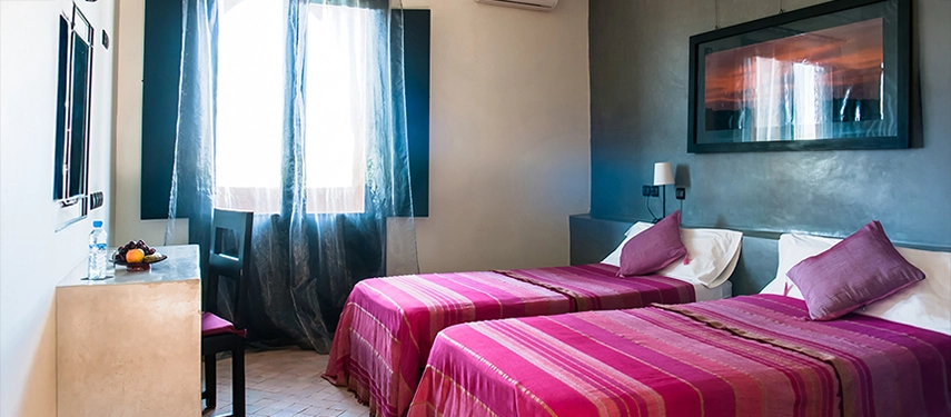 Twin room with purple-striped bedding, soft lighting, and desk with fruit and water.