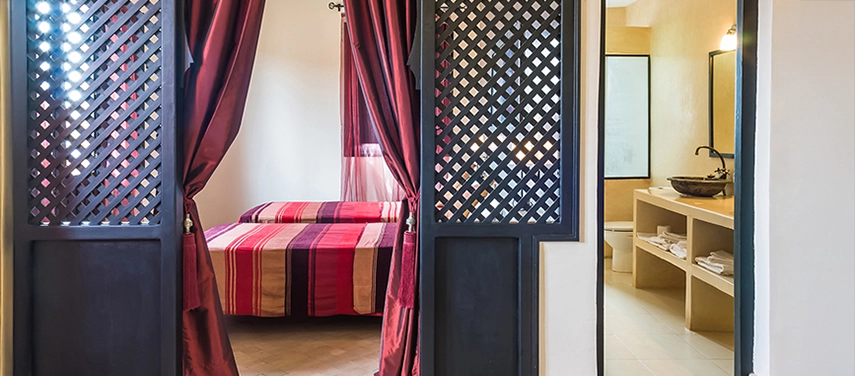 Bedroom entrance framed by carved wooden lattice doors, opening to a red-striped bed and en suite bathroom.