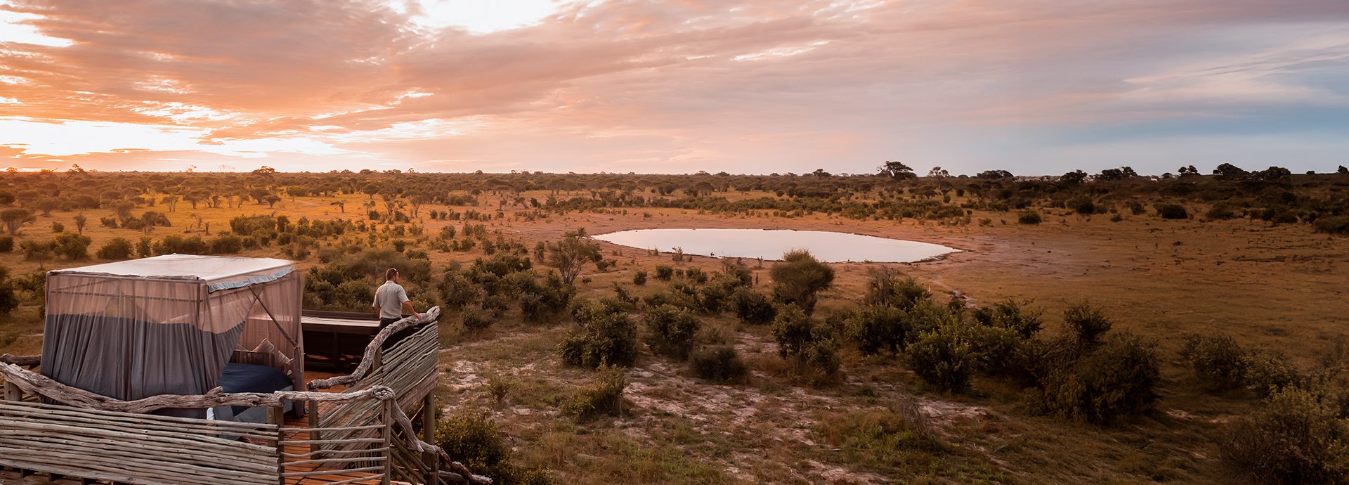 A guest stands beside a canopied Skybed platform, watching sunrise over the Khwai plains.