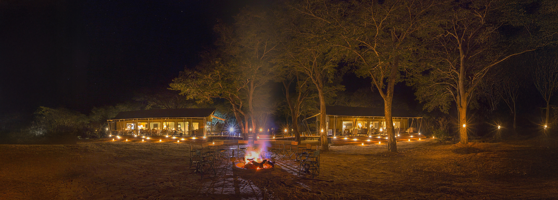 Night-time view of Verney’s Camp with glowing lanterns and a central campfire between the main tents.