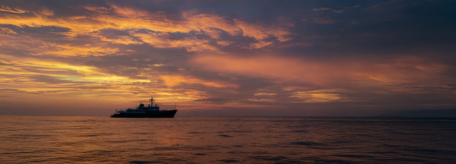 Aqua Blu yacht silhouetted against a vivid sunset sky, its reflection shimmering across calm Indonesian waters.