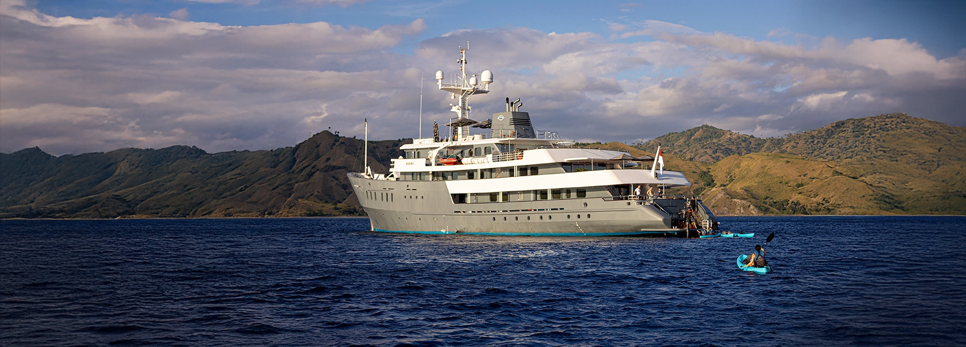 A kayaker paddles near Aqua Blu anchored in clear blue seas, with rugged Indonesian islands rising in the background.