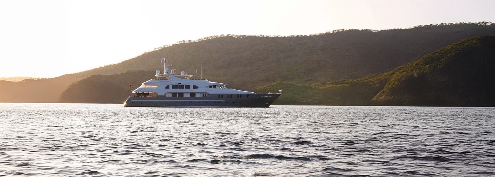 Aqua Mare sails along the Galápagos coastline, framed by soft morning light and emerald volcanic hills in the background.
