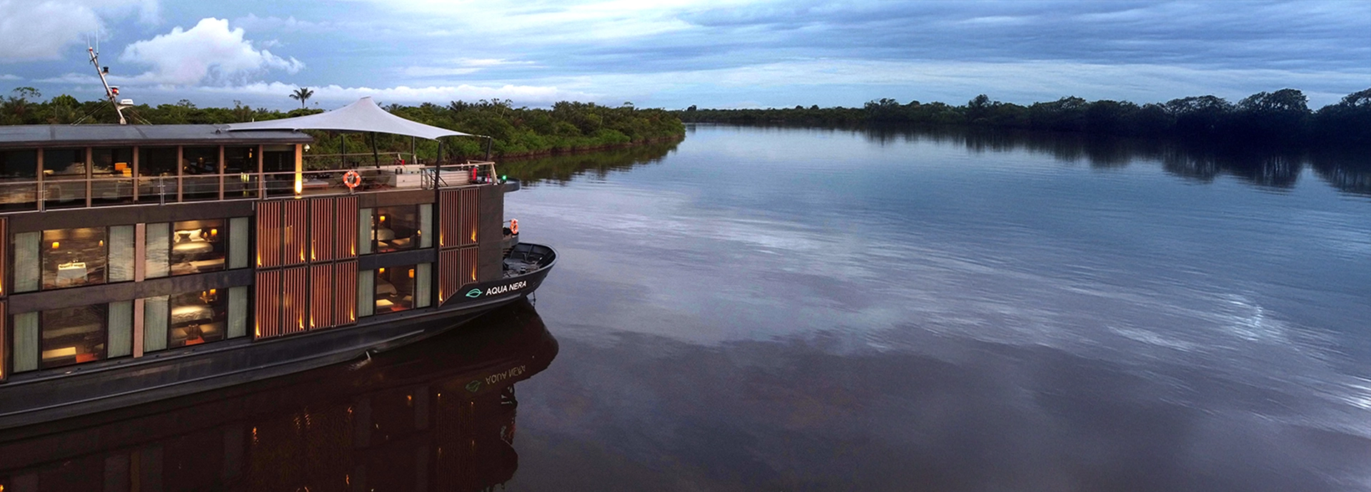 Aqua Nera gliding along the Amazon River at dusk, its sleek dark exterior reflecting soft golden light on the tranquil water.