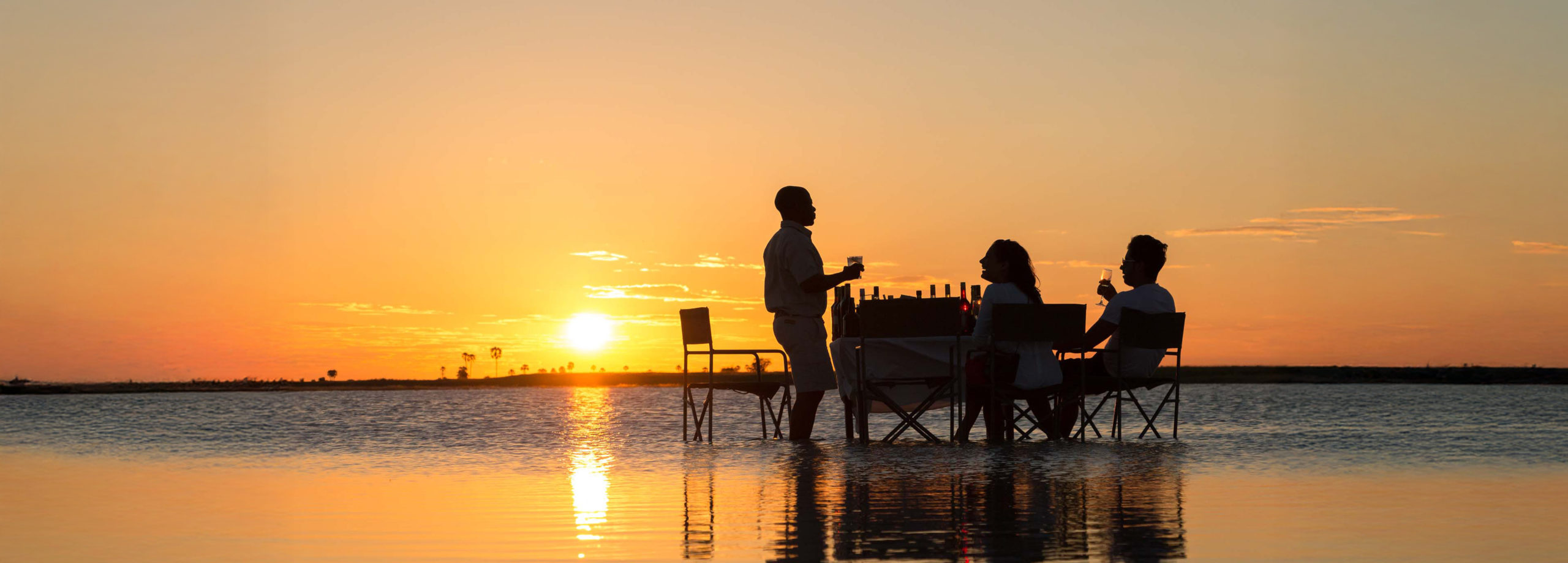 A guide serves sundowner drinks to guests on the reflective surface of the salt pans at sunset.