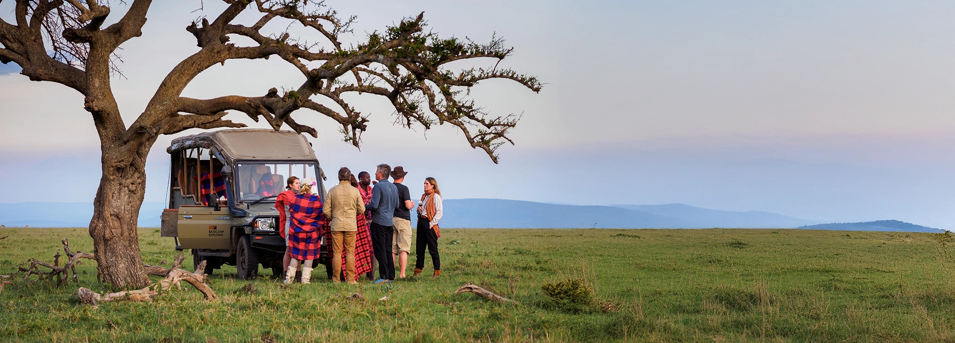 Wide view of a safari group interacting with Maasai guides under a sprawling acacia tree, with Basecamp Masai Mara’s vehicle and distant hills in the background.