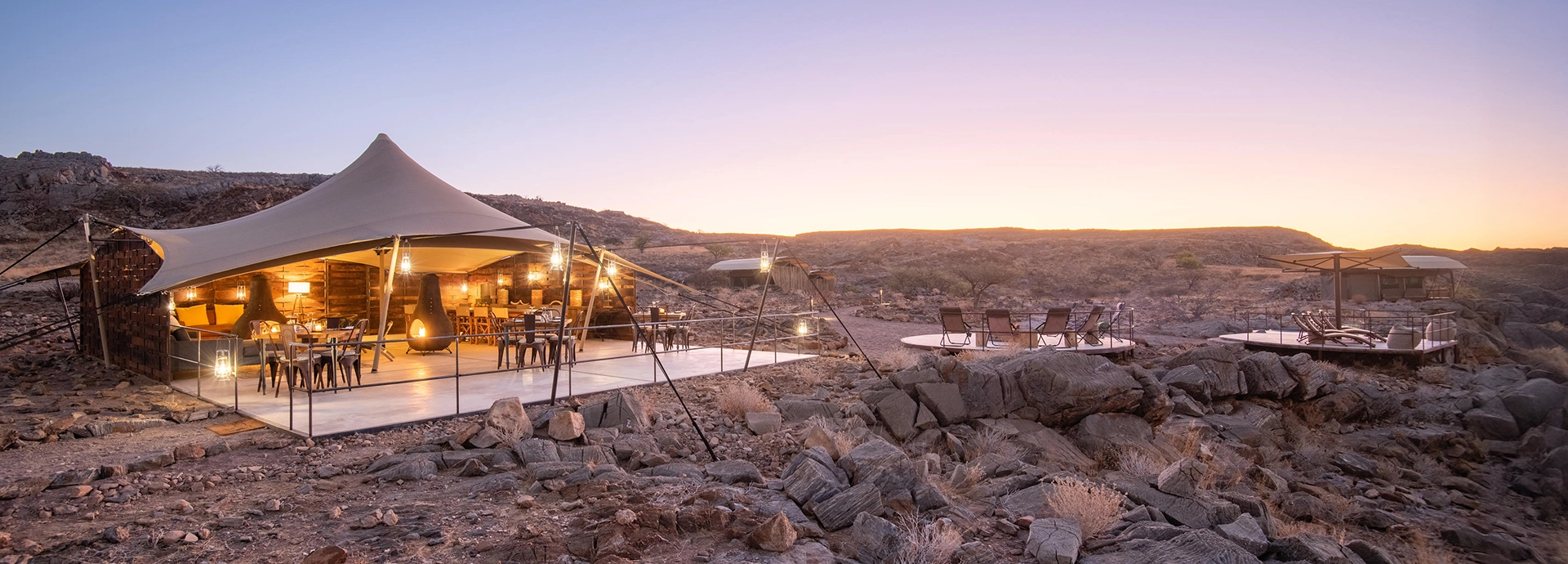 The main dining and lounge area at Camp Doros illuminated at dusk, blending canvas, wood, and stone in a rugged desert landscape.