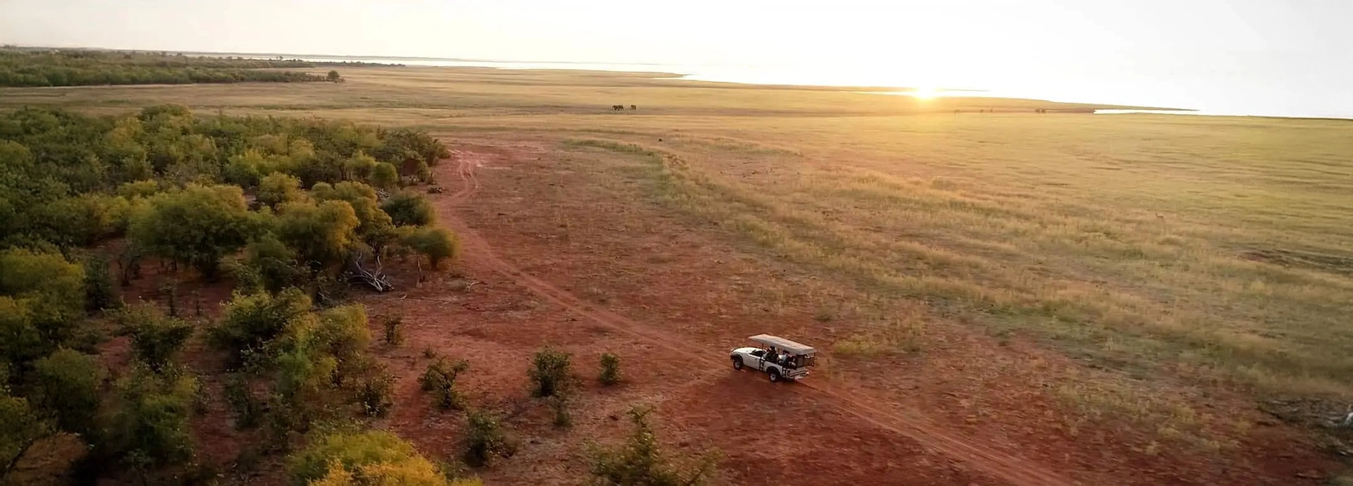 Aerial view of a safari vehicle crossing open plains near Lake Kariba during golden hour, showcasing the vast landscape around Changa Safari Camp.