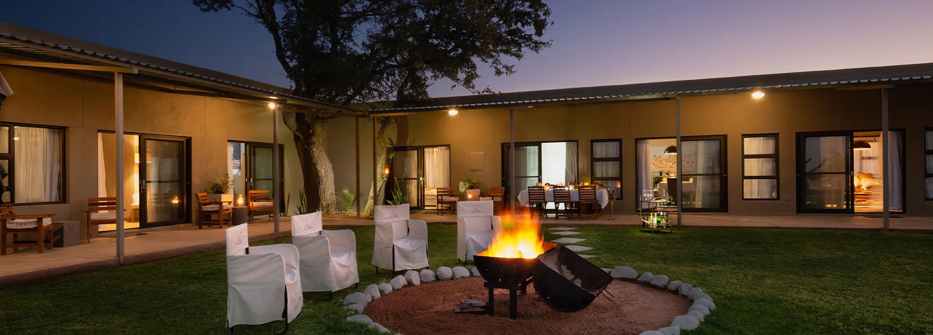 Outdoor firepit area with chairs and dining table at Safari House as evening lights glow.