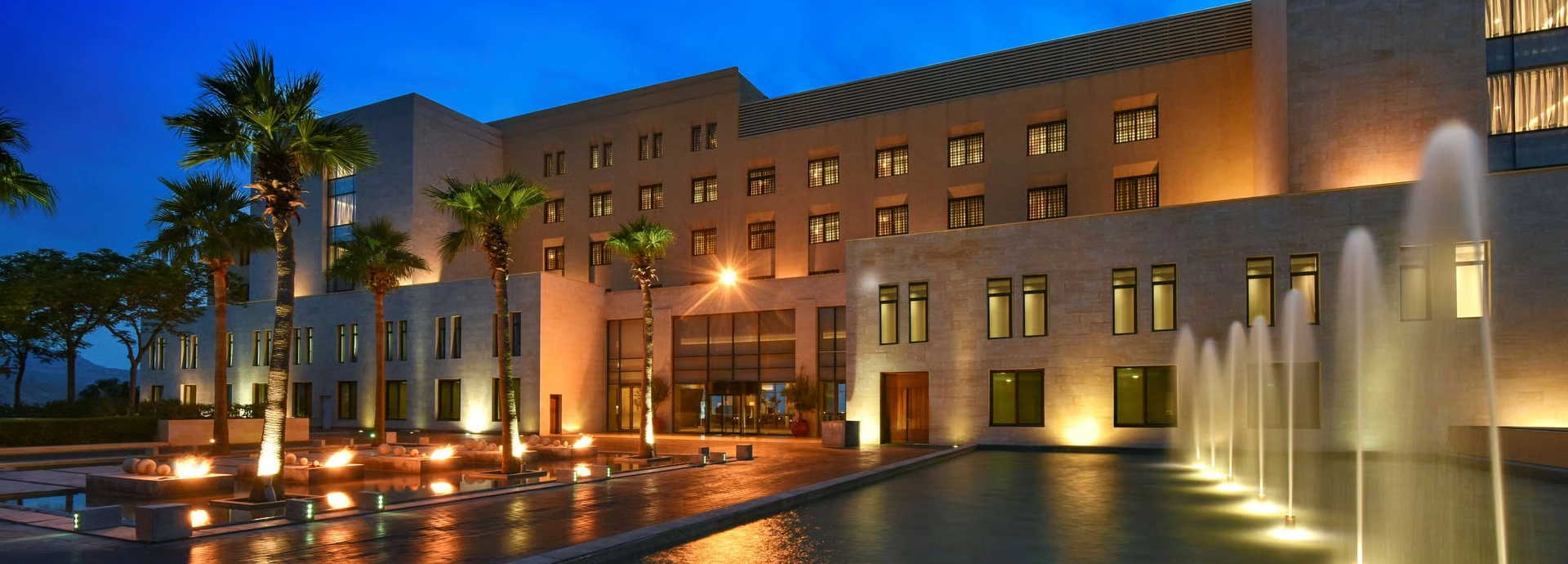 The illuminated main building of Kempinski Hotel Ishtar Dead Sea with palm-lined water features and a serene evening ambience.