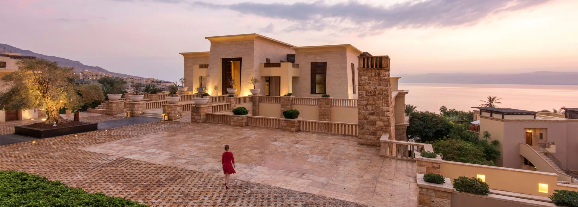 Grand stone terraces and palace-style architecture overlooking the Dead Sea at sunset, with warm lighting and expansive open courtyards.
