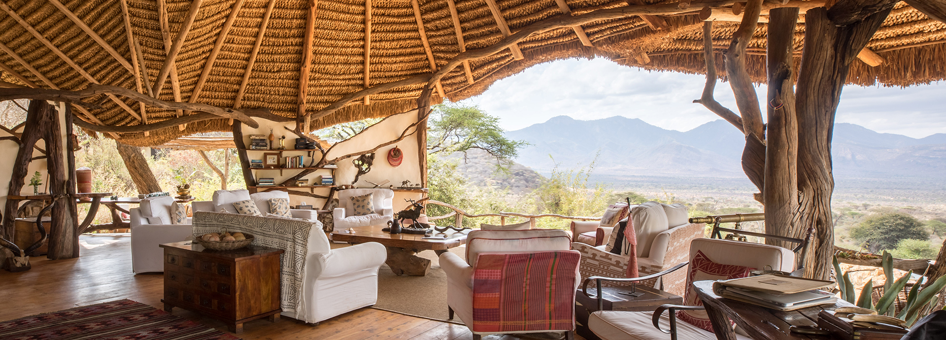 A cosy open-plan lounge at Sarara Camp with rustic wooden beams, white armchairs, and panoramic views of the Mathews Range.