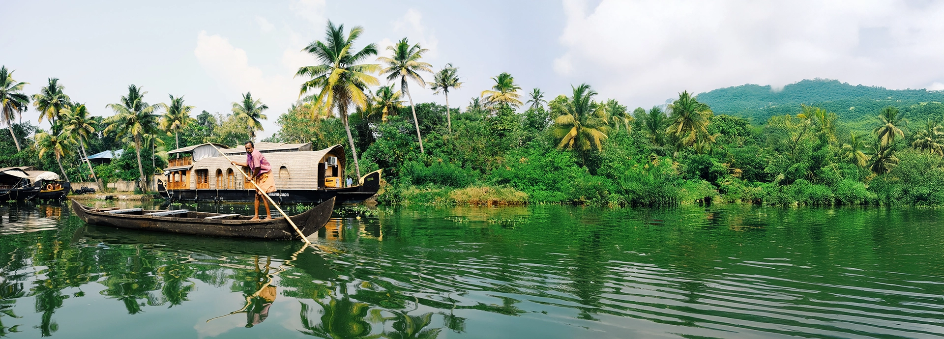 A traditional wooden canoe glides through Kerala’s backwaters, with palm-lined banks and a Kettuvallam reflecting on the still water.