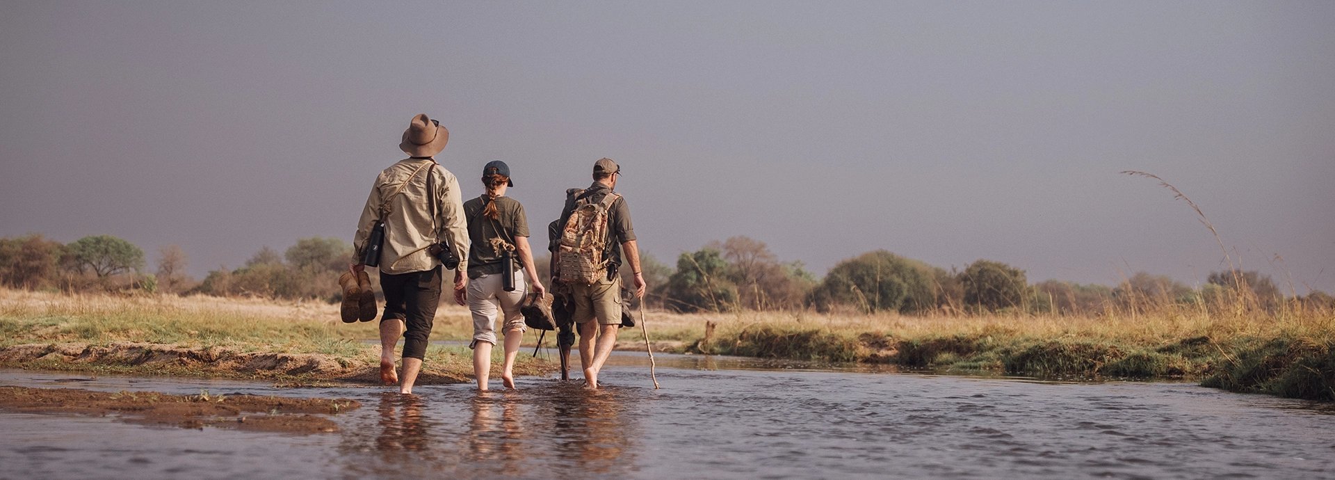 Guests and guides wade barefoot across a shallow river on a walking safari, carrying boots and equipment through pristine wilderness.