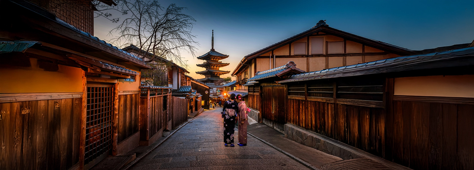 A lantern-lit street in Kyoto’s historic district at dusk, where traditional wooden townhouses frame a quiet evening stroll.