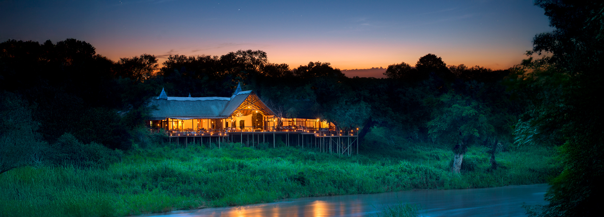 A twilight view of Lion Sands Narina Lodge glowing warmly above the Sabie River.