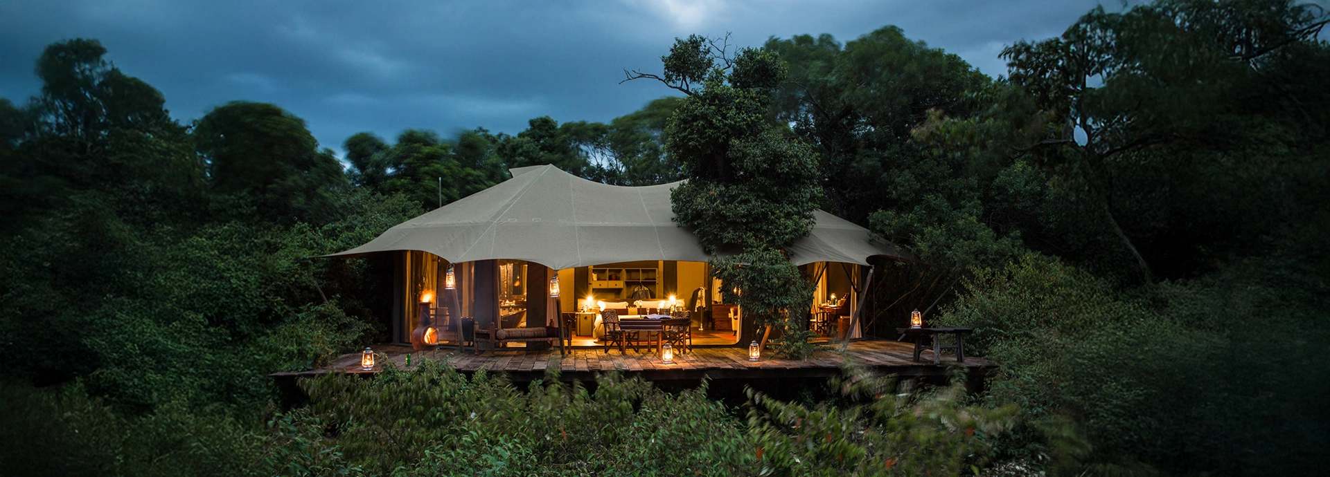 Evening view of a Mara Plains suite in Kenya, glowing softly under canvas with wooden decks suspended above lush riverbank vegetation.