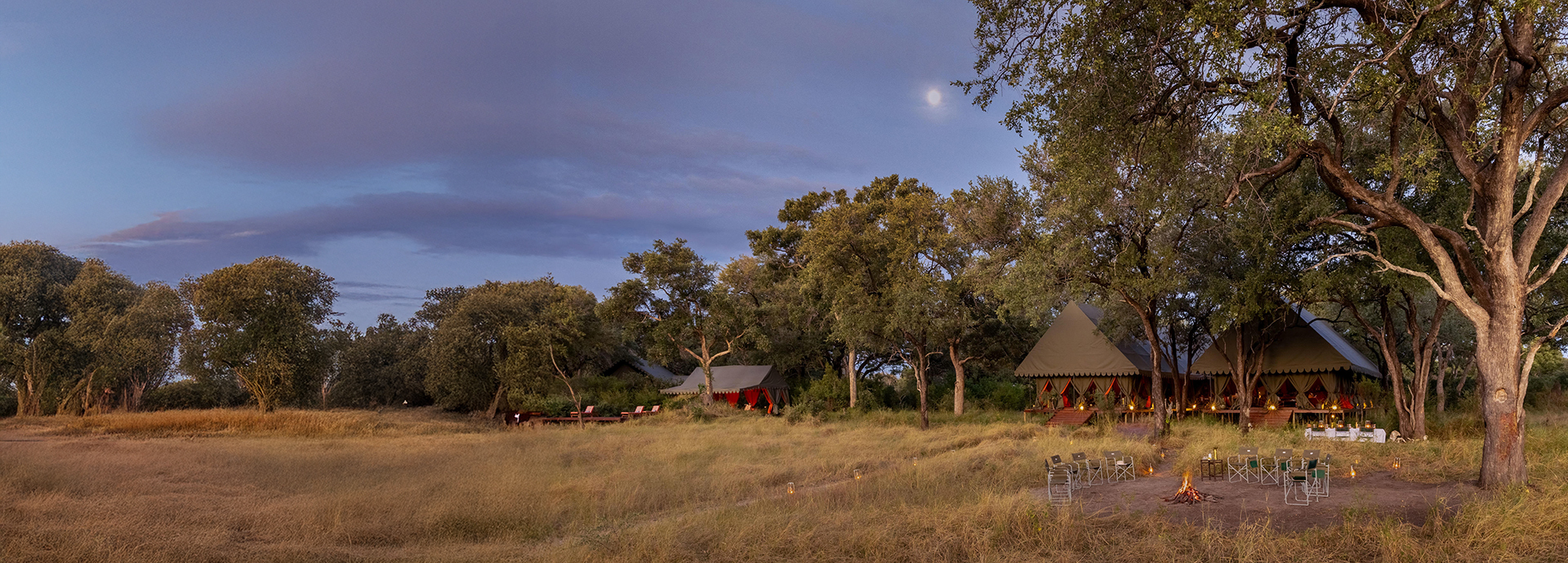 Panoramic view of Mbamba Camp, with tents nestled under trees and a firepit set in the grassland.