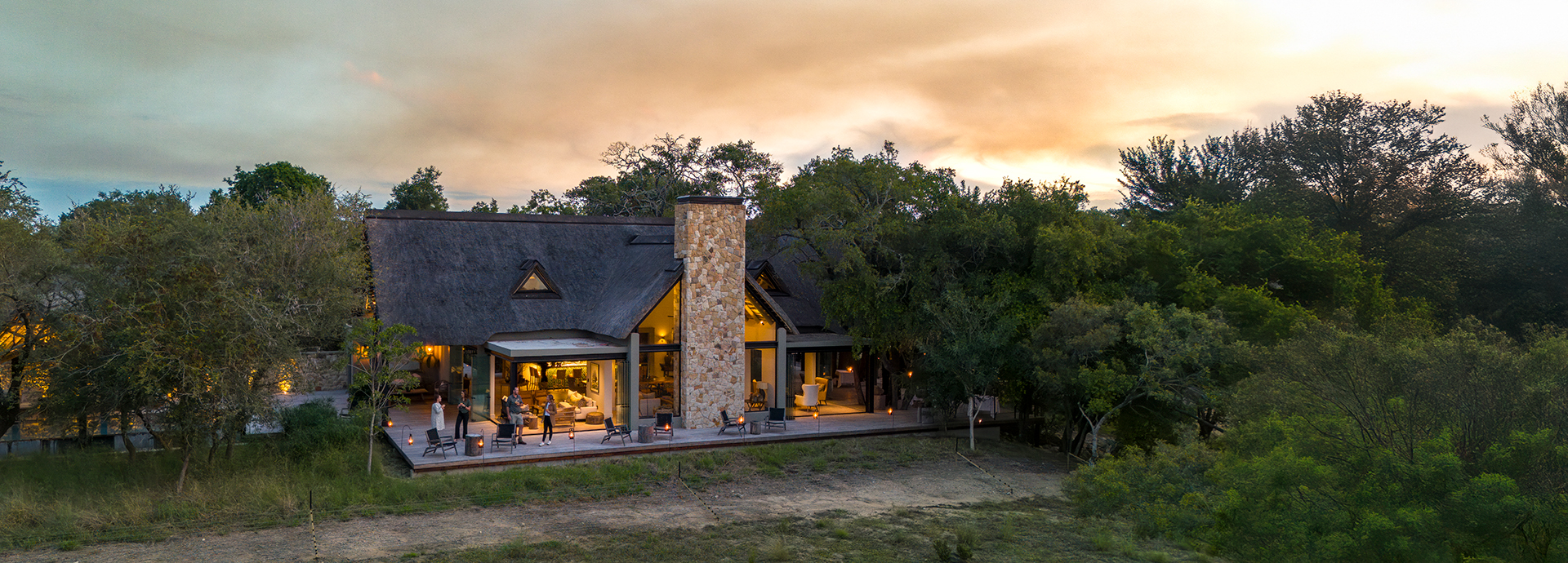 A view of Monwana Lodge at sunset, with glowing interiors and a thatched roof surrounded by bushland.